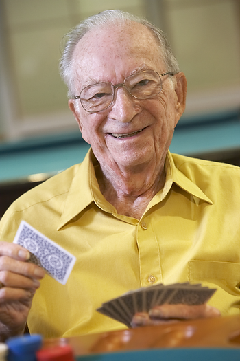 An elderly man in a yellow shirt is holding a playing card and smiling.