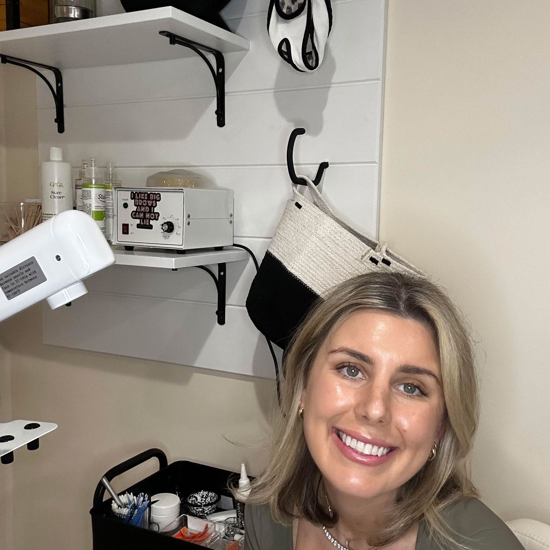 Woman smiles, stands in front of a shelf with beauty products, a waxing machine, and a small black cart.