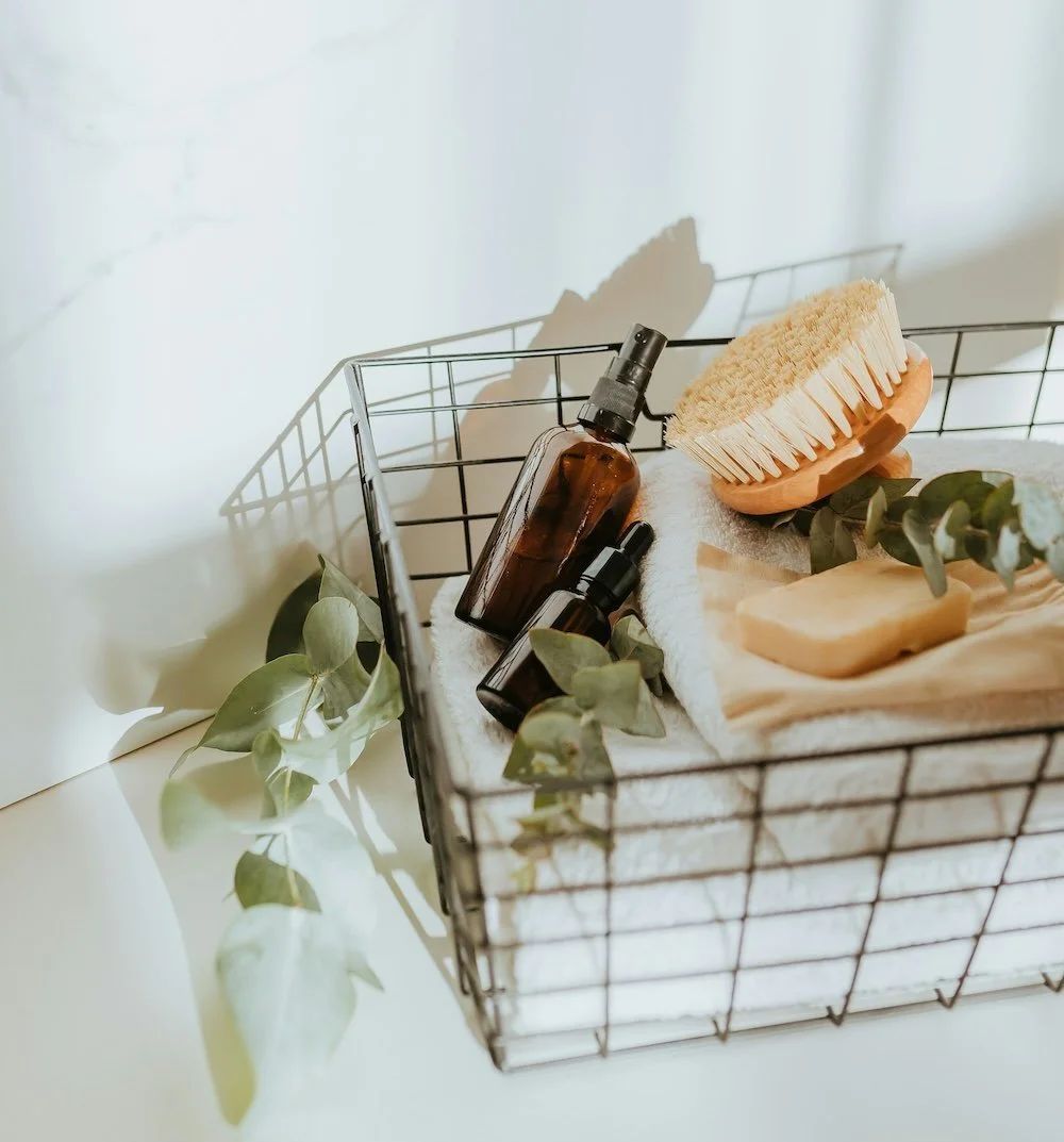 Wire basket with beauty products: brown bottles, a brush, soap, and greenery.
