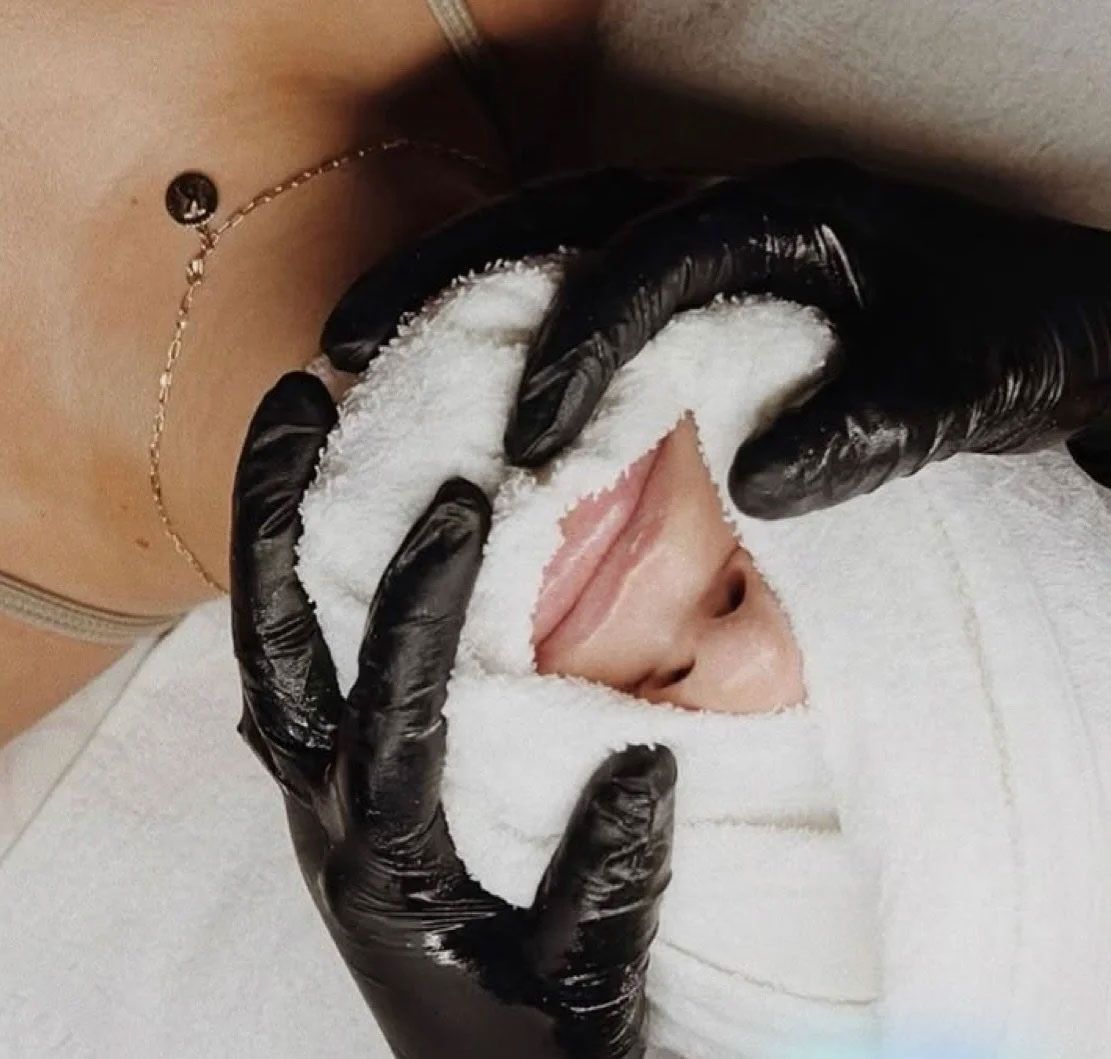 Hands in black gloves holding a white towel to a person's face during a facial treatment.