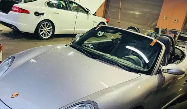 Silver Porsche Boxster in a garage, with a white Jaguar in the background.