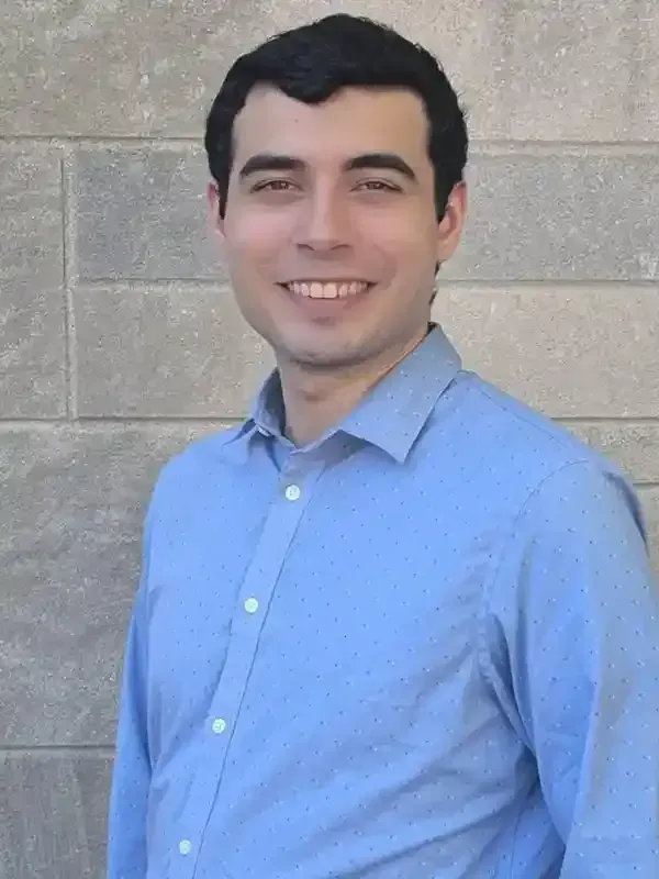 Man in a blue button-down shirt smiling in front of a gray brick wall.