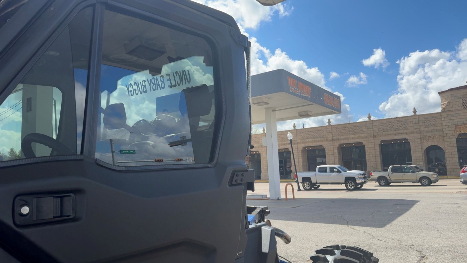 A view from inside a dark vehicle cabin looking out at a gas station with two parked pickup trucks under a blue sky.