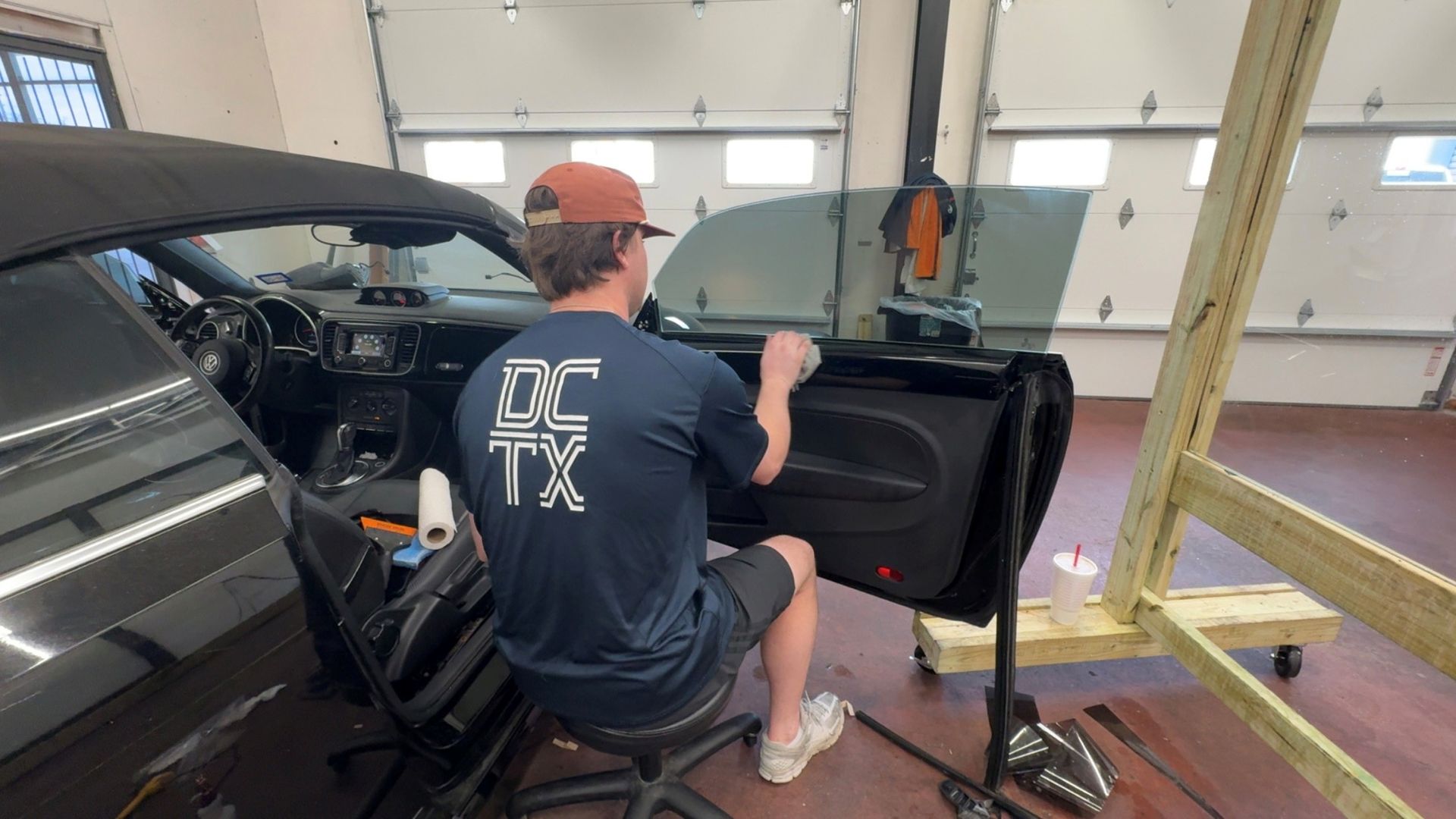 A technician wearing a navy shirt and brown cap sits on a stool, detailing the interior of a black car door in a garage.