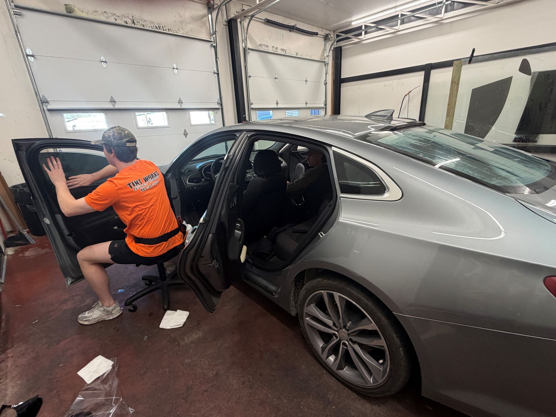 A technician in an orange shirt sits on a rolling chair, applying window tint to the rear driver-side window of a car.