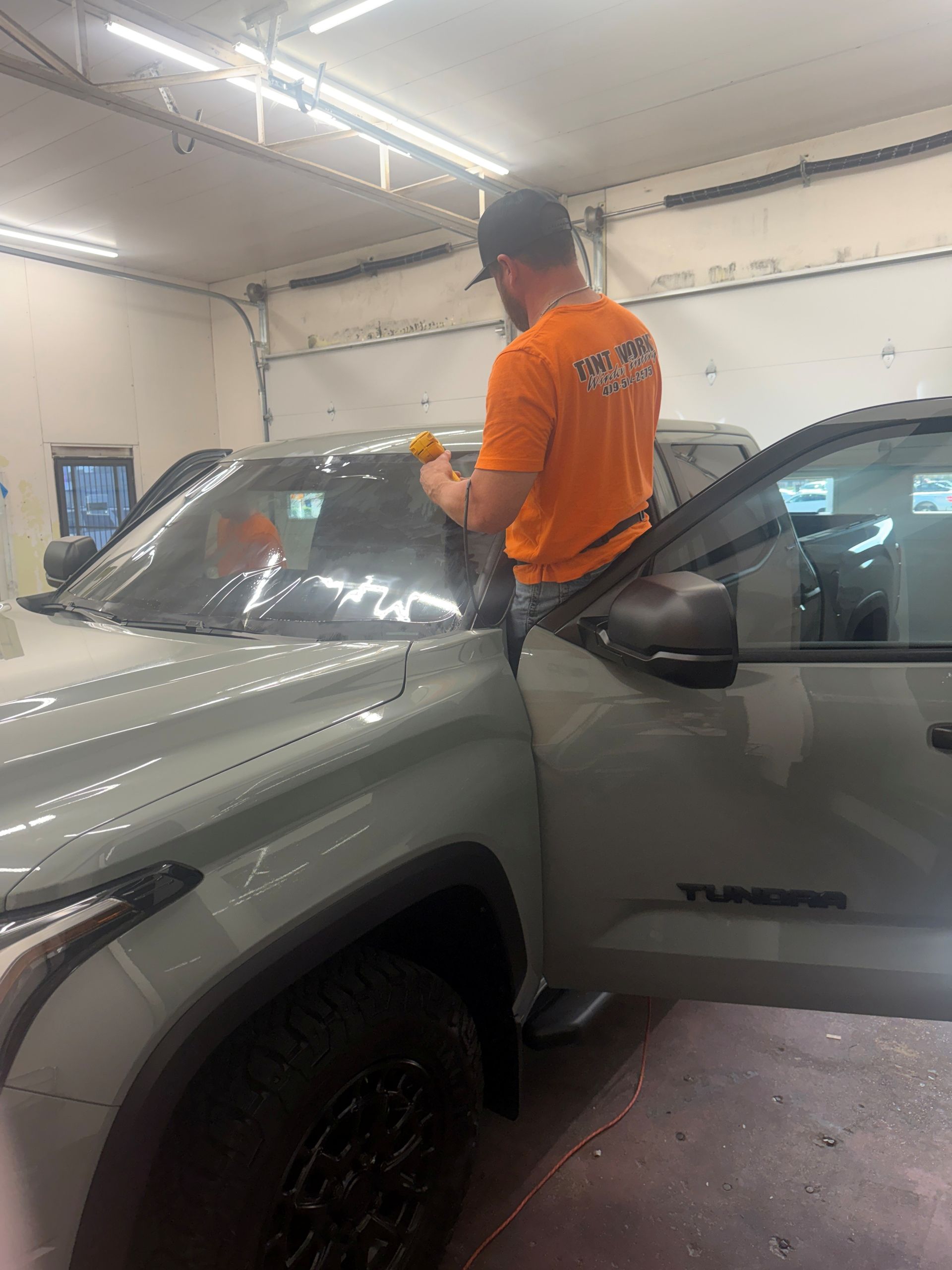 A person in an orange shirt works on applying window tint to the windshield of a grey Toyota Tundra in a garage.