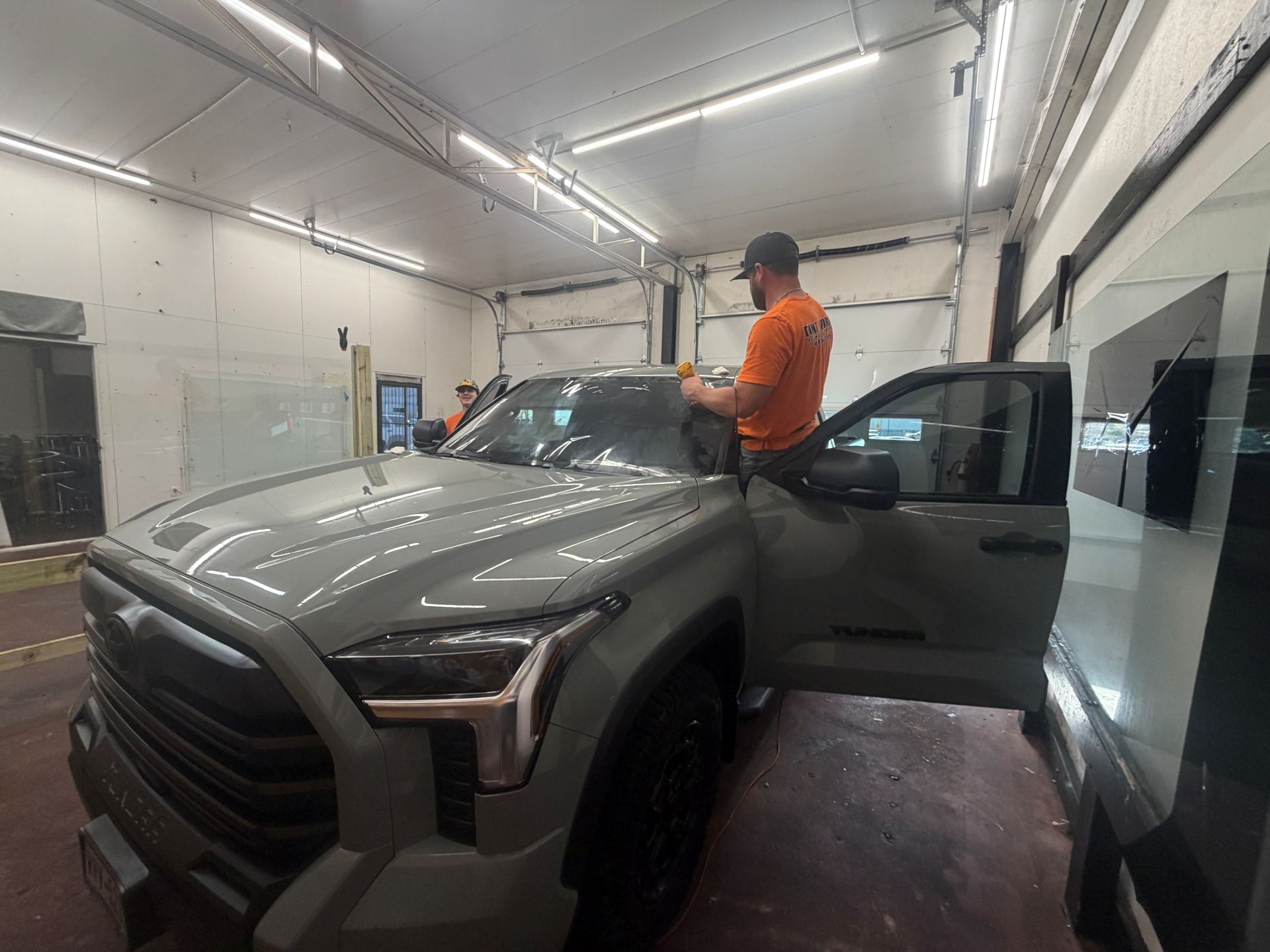 A person in an orange shirt works on the interior of a grey pickup truck inside a brightly lit automotive workshop.