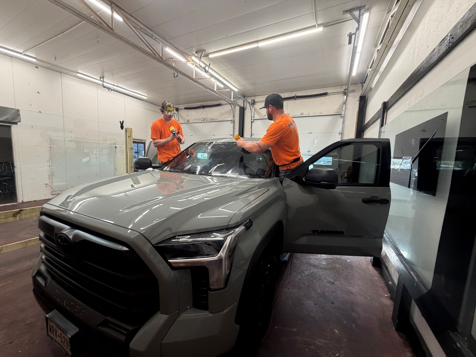 Two technicians in orange shirts work on installing window tint on a grey pickup truck inside a service garage.