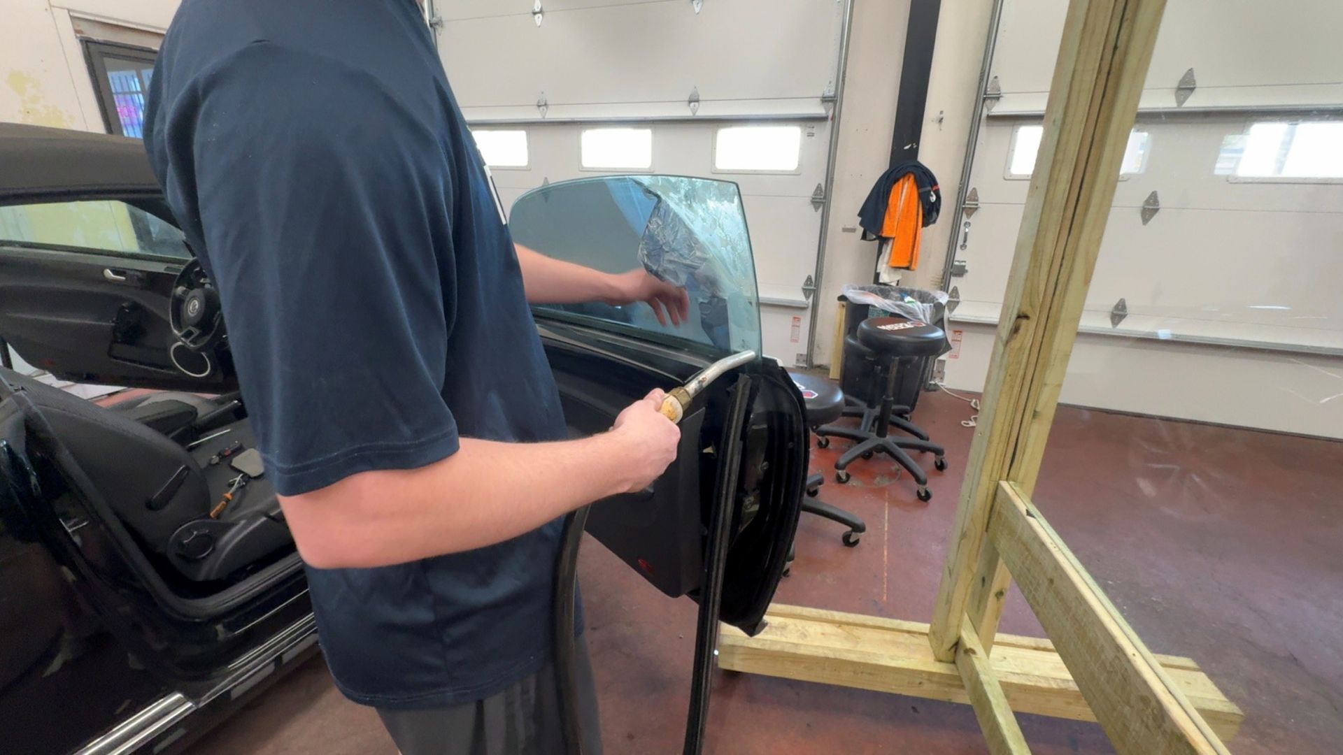 A person in a blue shirt uses a handheld tool to apply or smooth window tint on a car door in a garage.