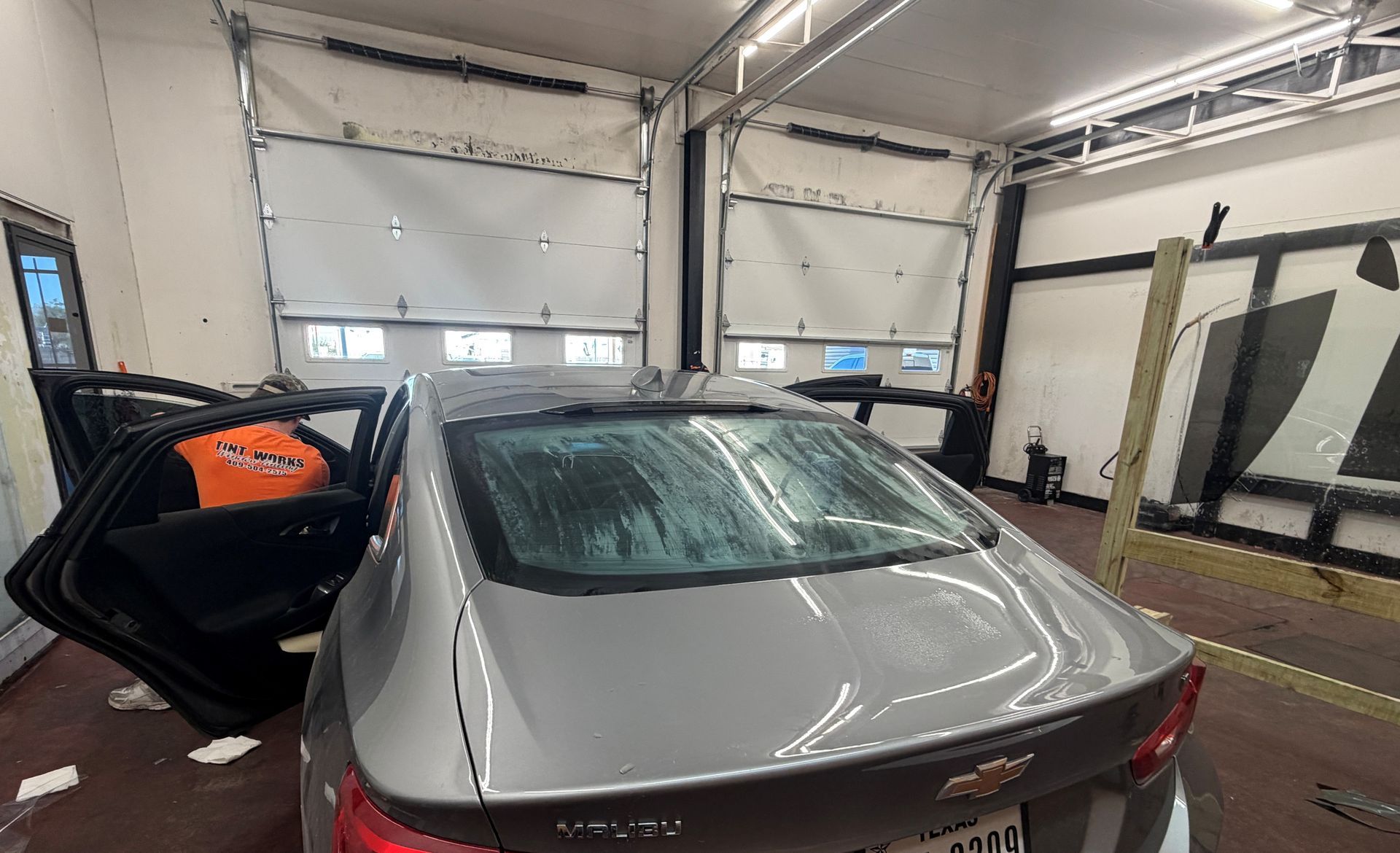 A silver Chevrolet Malibu parked in an auto repair shop with both rear doors open and a worker inside the vehicle.