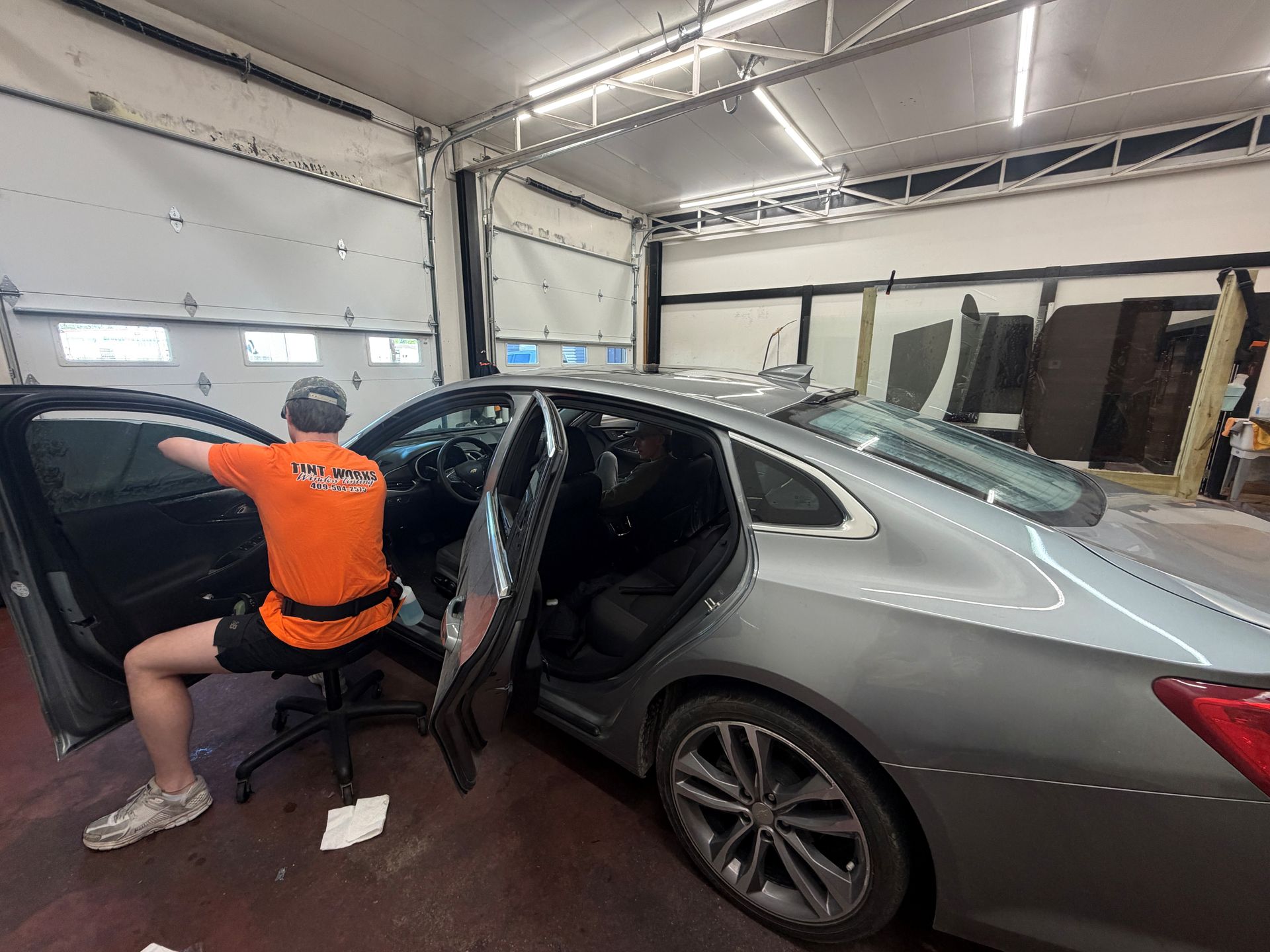 A person in an orange work shirt sits on a rolling stool, installing window tint on a grey car inside a garage.