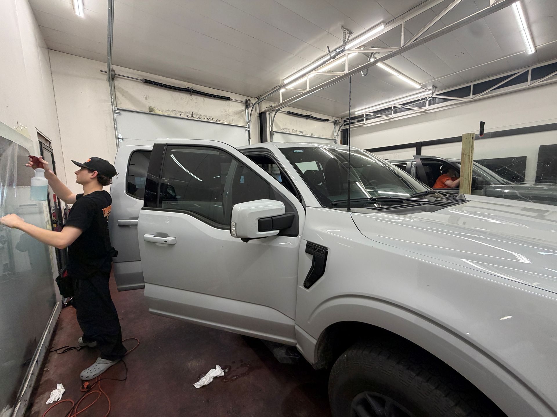 A technician sprays liquid on a glass surface in a garage while a white pickup truck sits in the foreground.