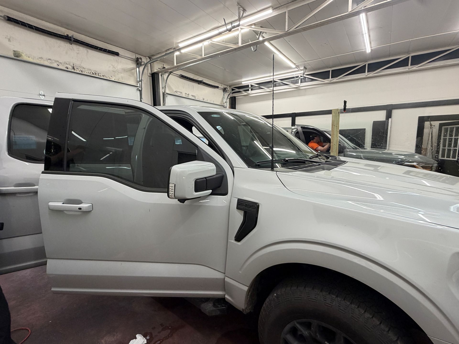 A white pickup truck with its driver-side door open inside a brightly lit auto body repair shop.