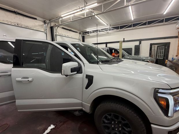 A light-colored Ford F-150 truck with its driver-side door open inside a well-lit auto shop.