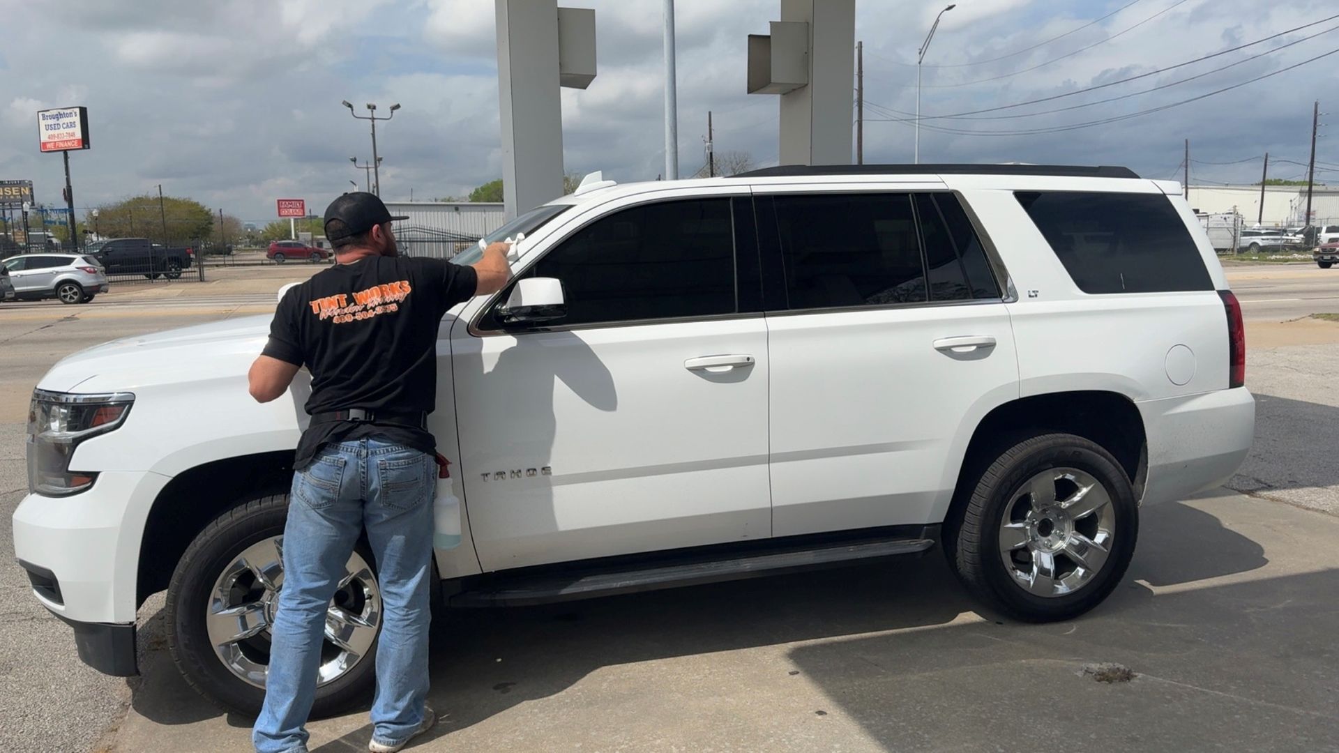 A person in a black t-shirt and jeans applies window tint to a white SUV parked at a gas station.