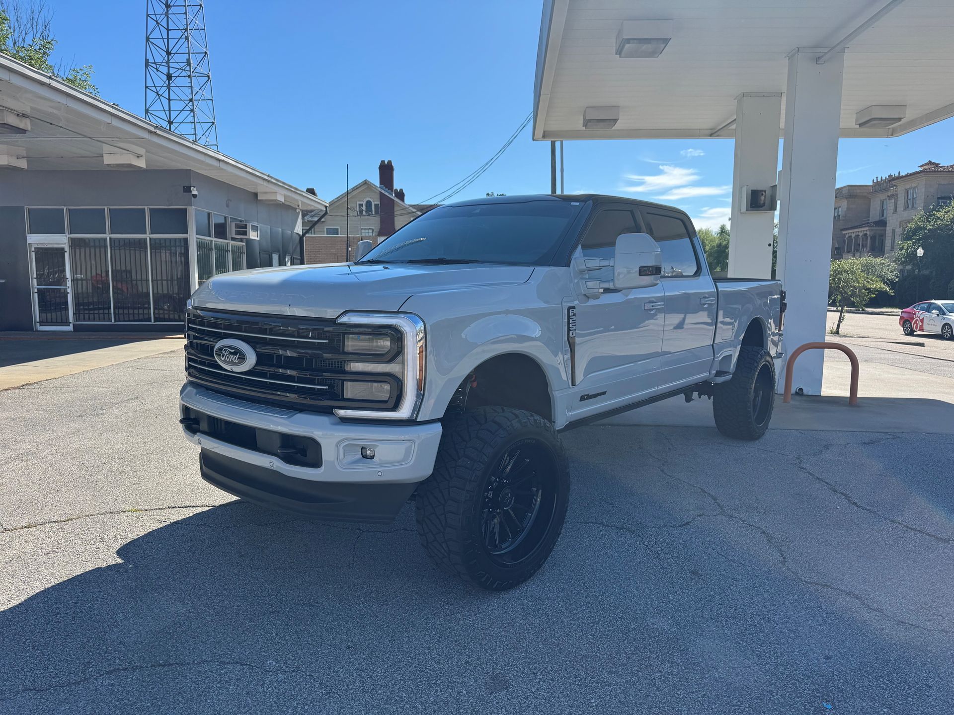 A light gray Ford Super Duty pickup truck with black wheels and a lift kit parked at a gas station on a sunny day.
