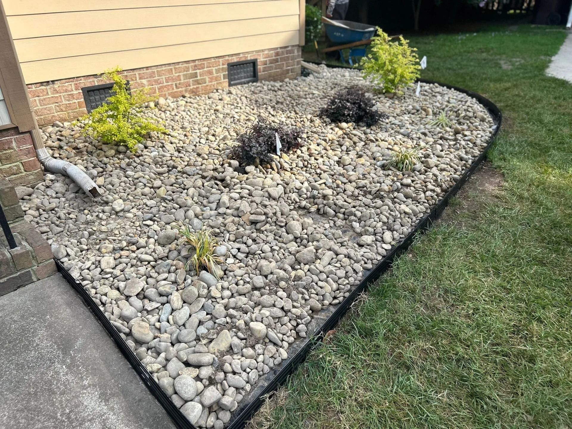 Rock-covered garden bed with green plants, bordered by black edging, next to a beige house and sidewalk.