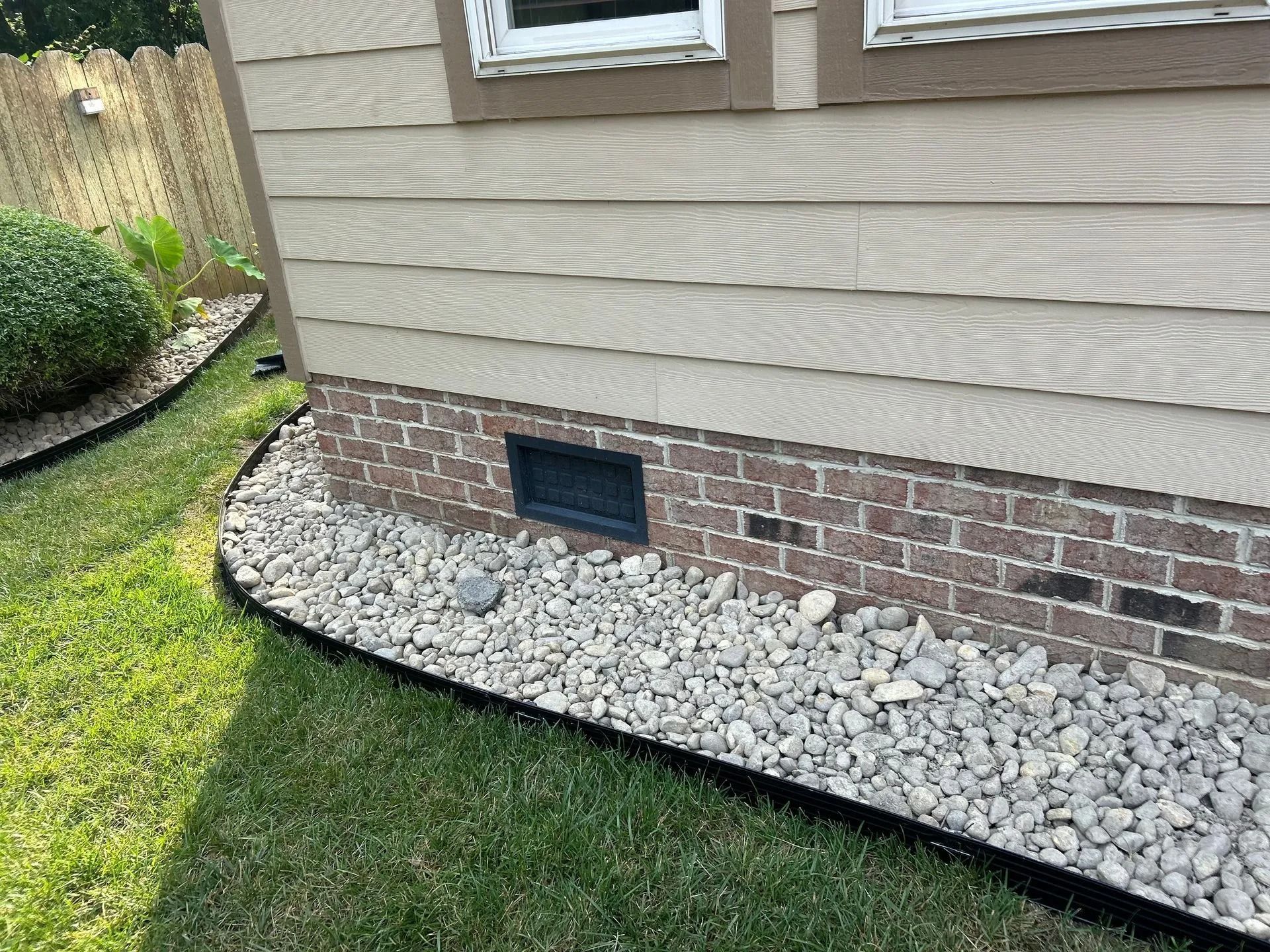 Corner of house with brick foundation, tan siding, and gravel bed next to grass.