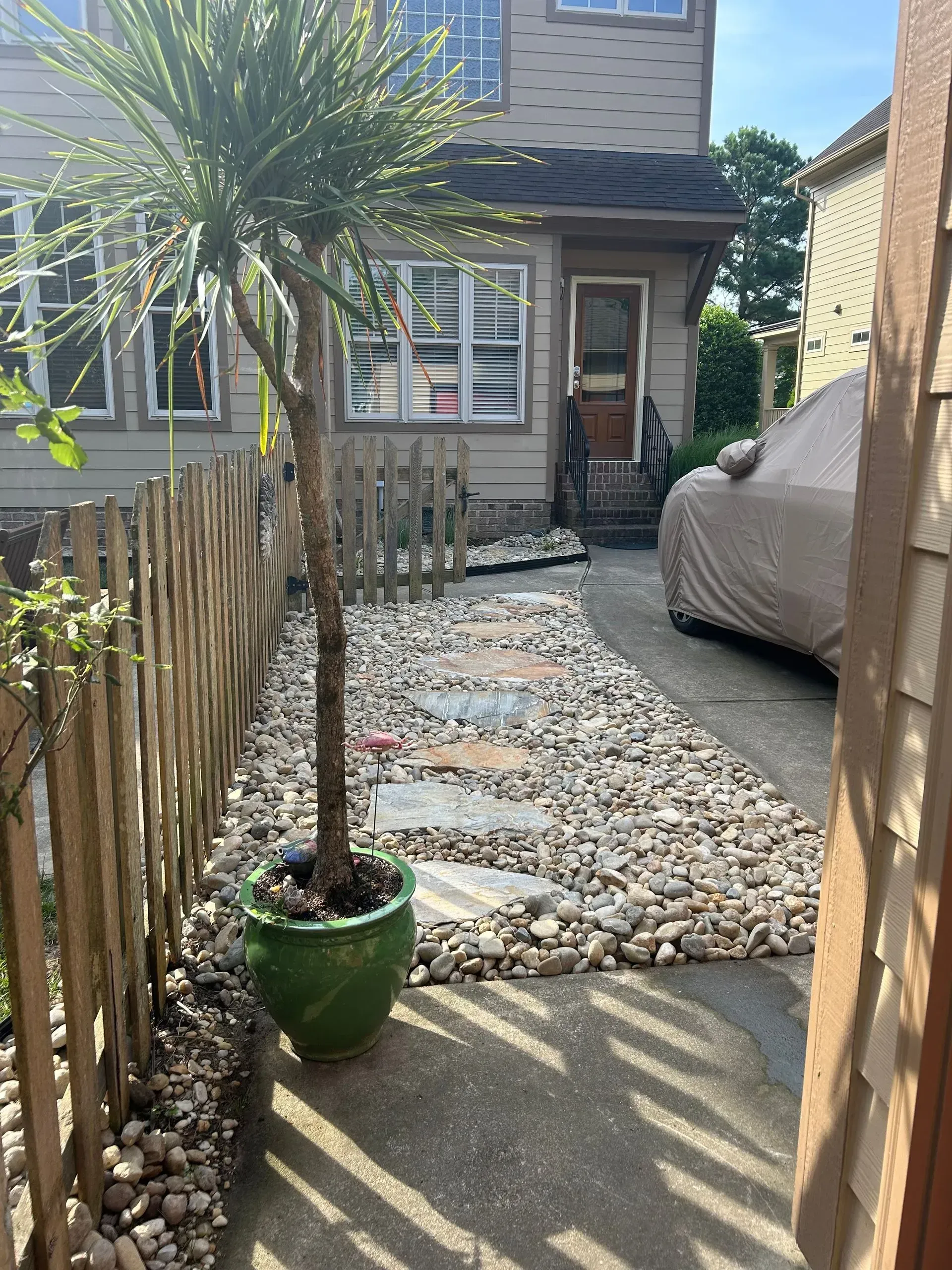 Stone path leads to a house with a car parked beside it; a tree in a green pot stands on the side.