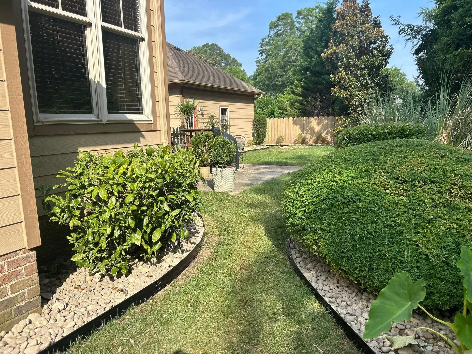 Lush green backyard with a grassy path flanked by rock borders, shrubs, and a partial view of a house.