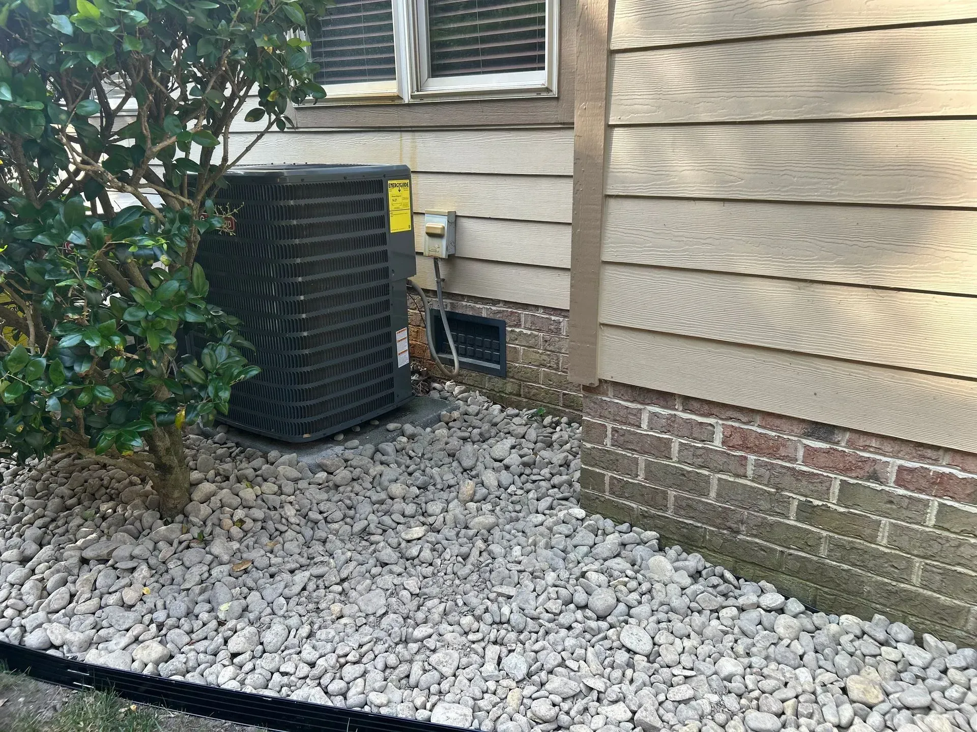 Air conditioning unit outside a house with gray siding and brick foundation, surrounded by gray gravel.