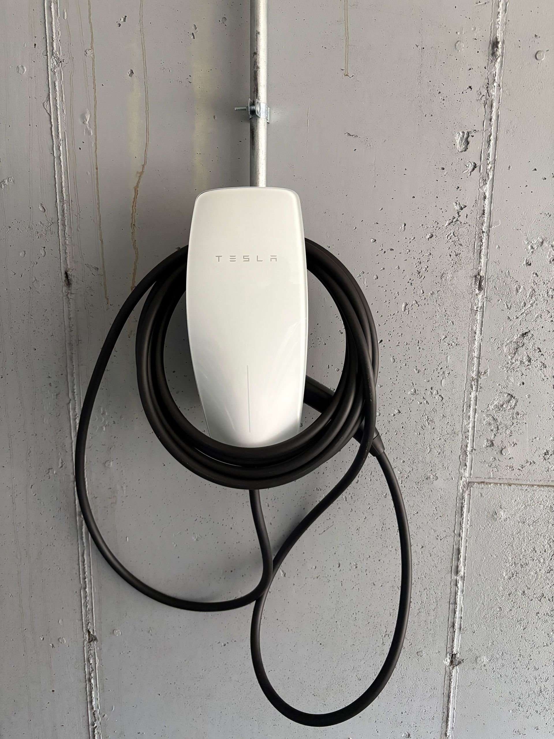 White Tesla wall charger with black cable coiled on a gray wall.