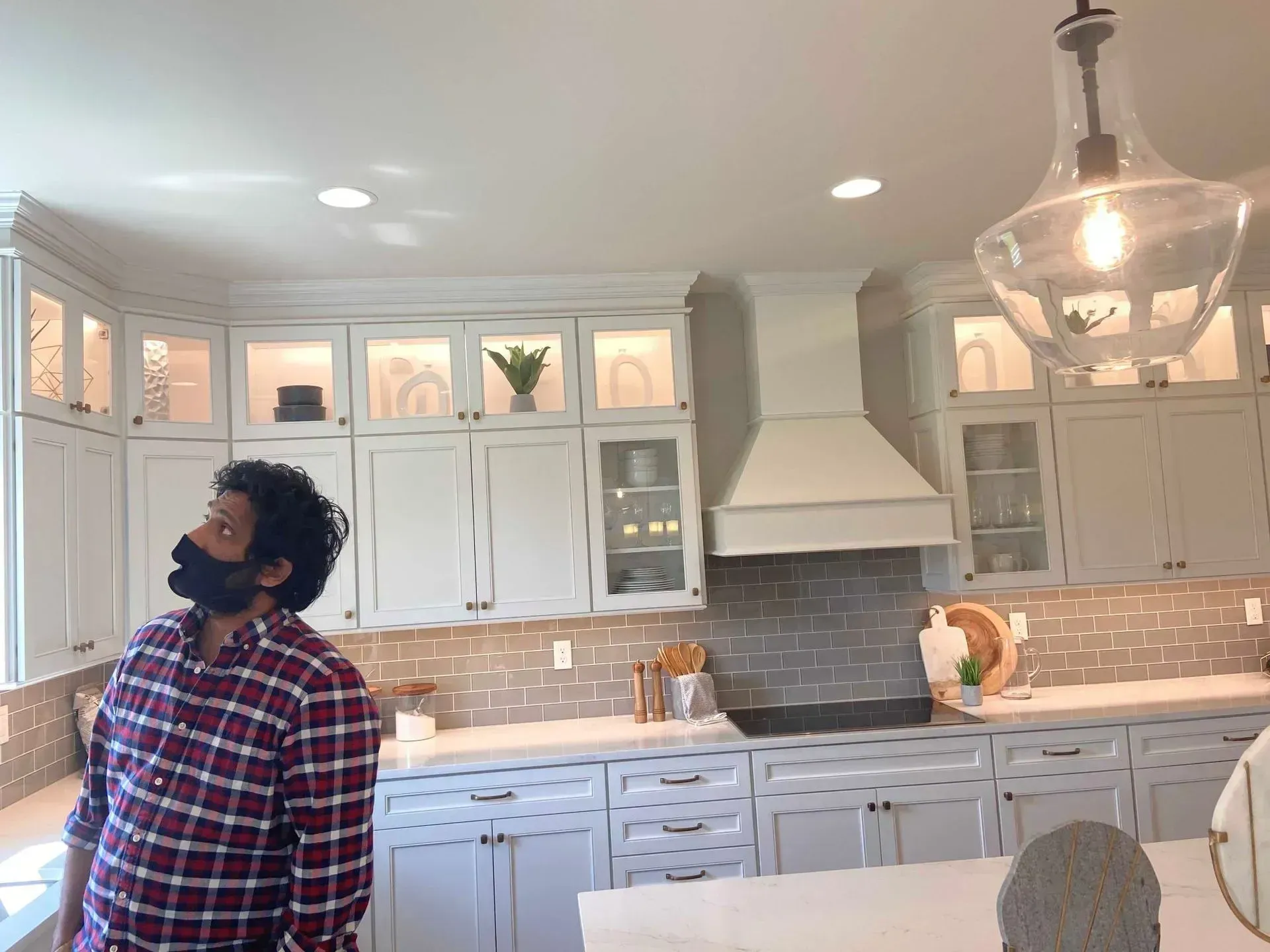 Man in a plaid shirt and mask in a modern kitchen, looking up at the ceiling lights