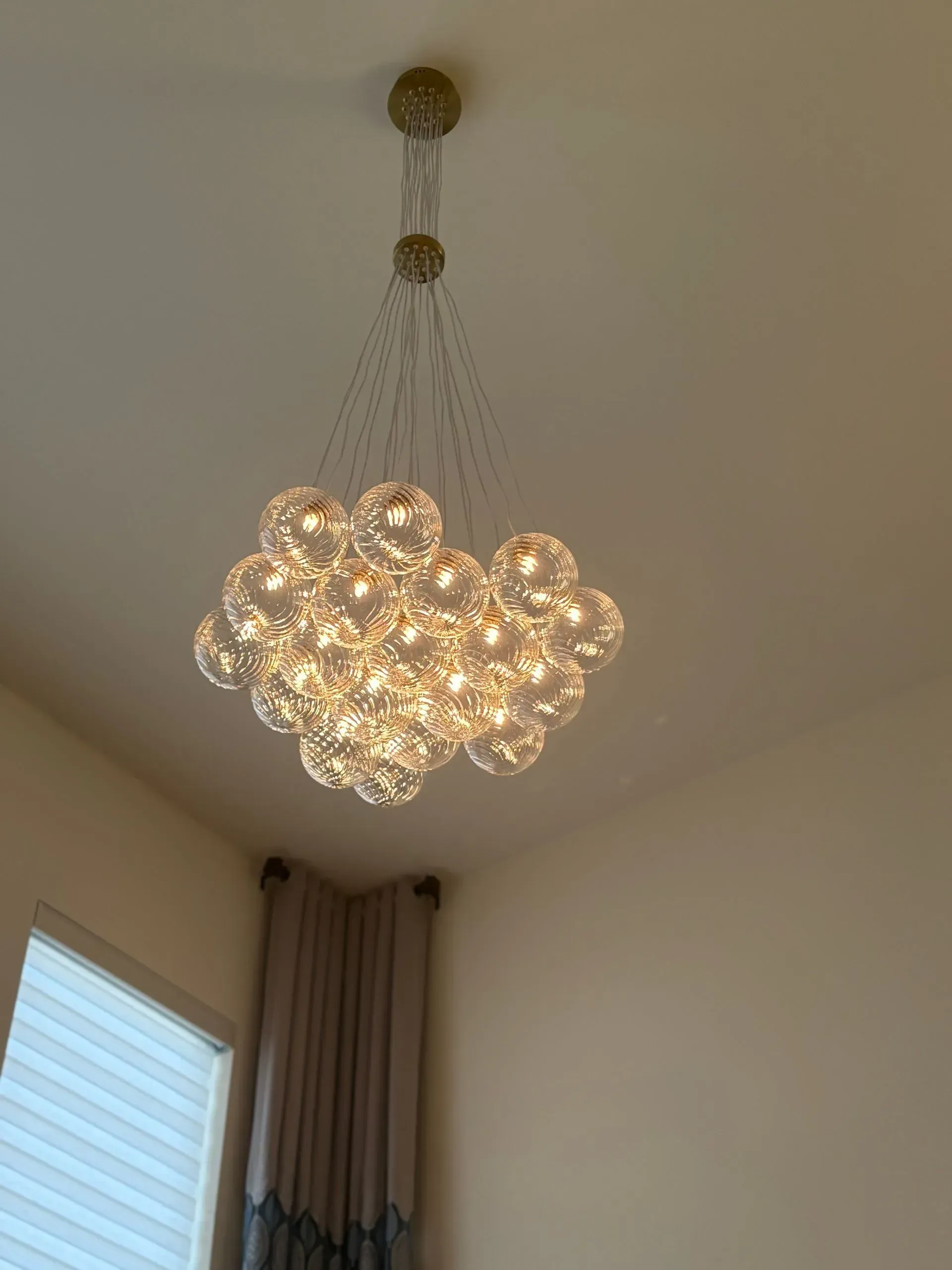 A modern chandelier with multiple illuminated glass globes hangs from a white ceiling near a window and curtains