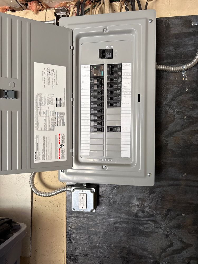 Gray electrical panel with open door, circuit breakers, and a conduit on a dark wall.