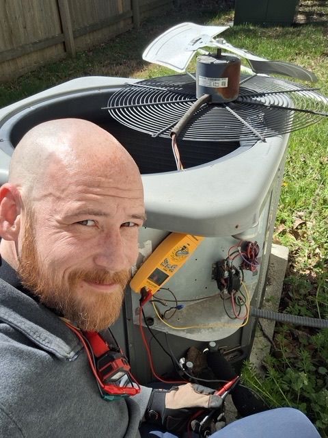 Bald man with a beard working on an air conditioning unit outside, using a multimeter.