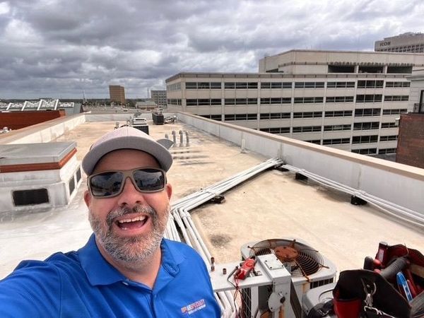 Man on a rooftop smiles at camera. Cloudy sky, commercial buildings. He wears a hat, sunglasses, and blue shirt.
