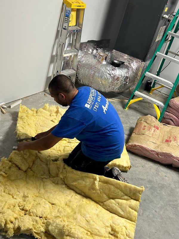 Man kneeling, cutting insulation on a concrete floor near ladders and ductwork.