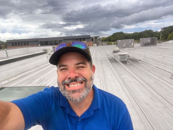 Man smiling on a metal roof, blue shirt, baseball cap, cloudy sky, commercial building in background.