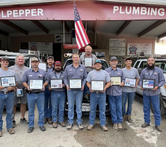 A group of plumbing technicians in uniform holding certificates stand in front of their shop under an American flag.