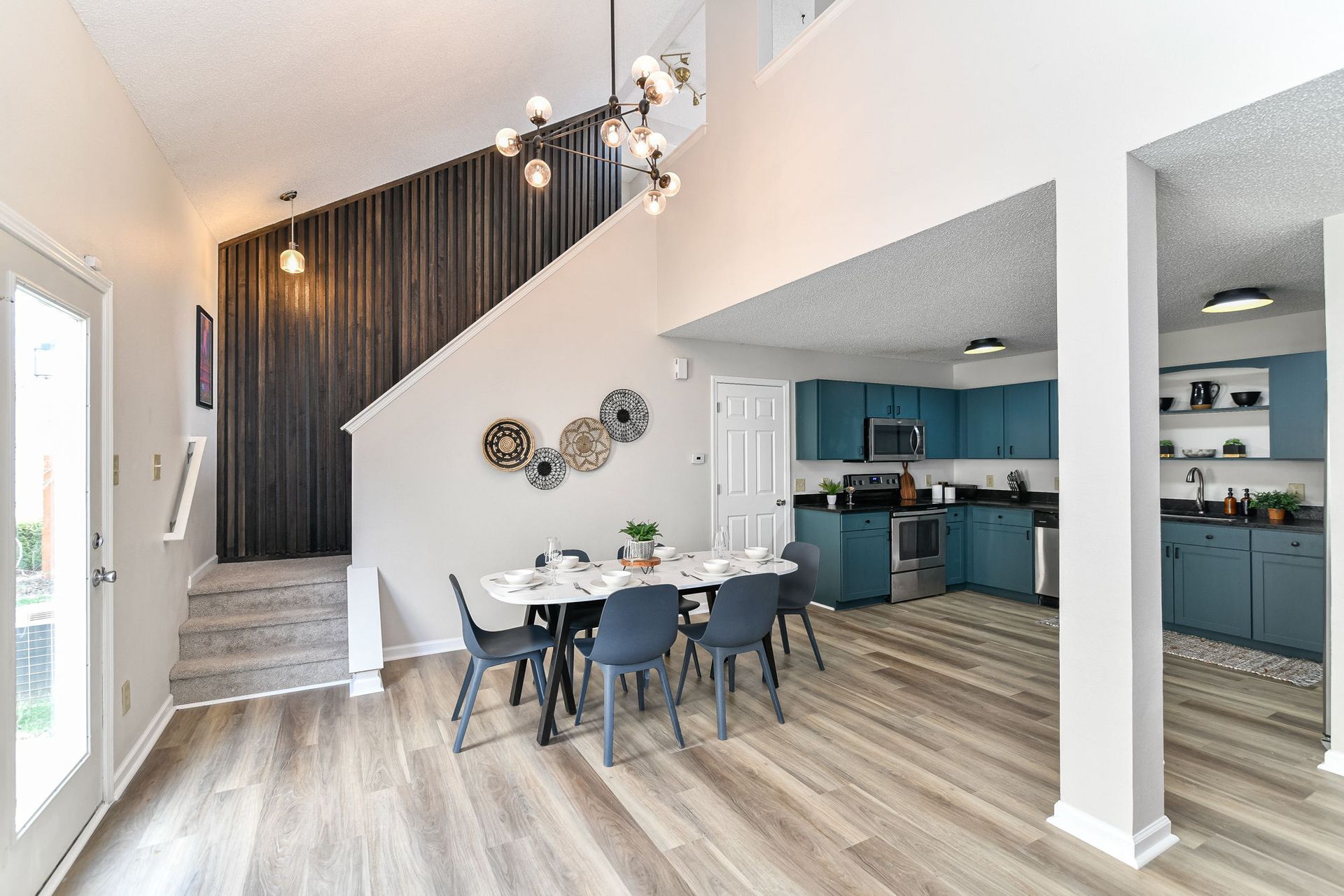 Spacious dining room with blue kitchen in the background. Light wood floors, high ceilings, and modern light fixtures.