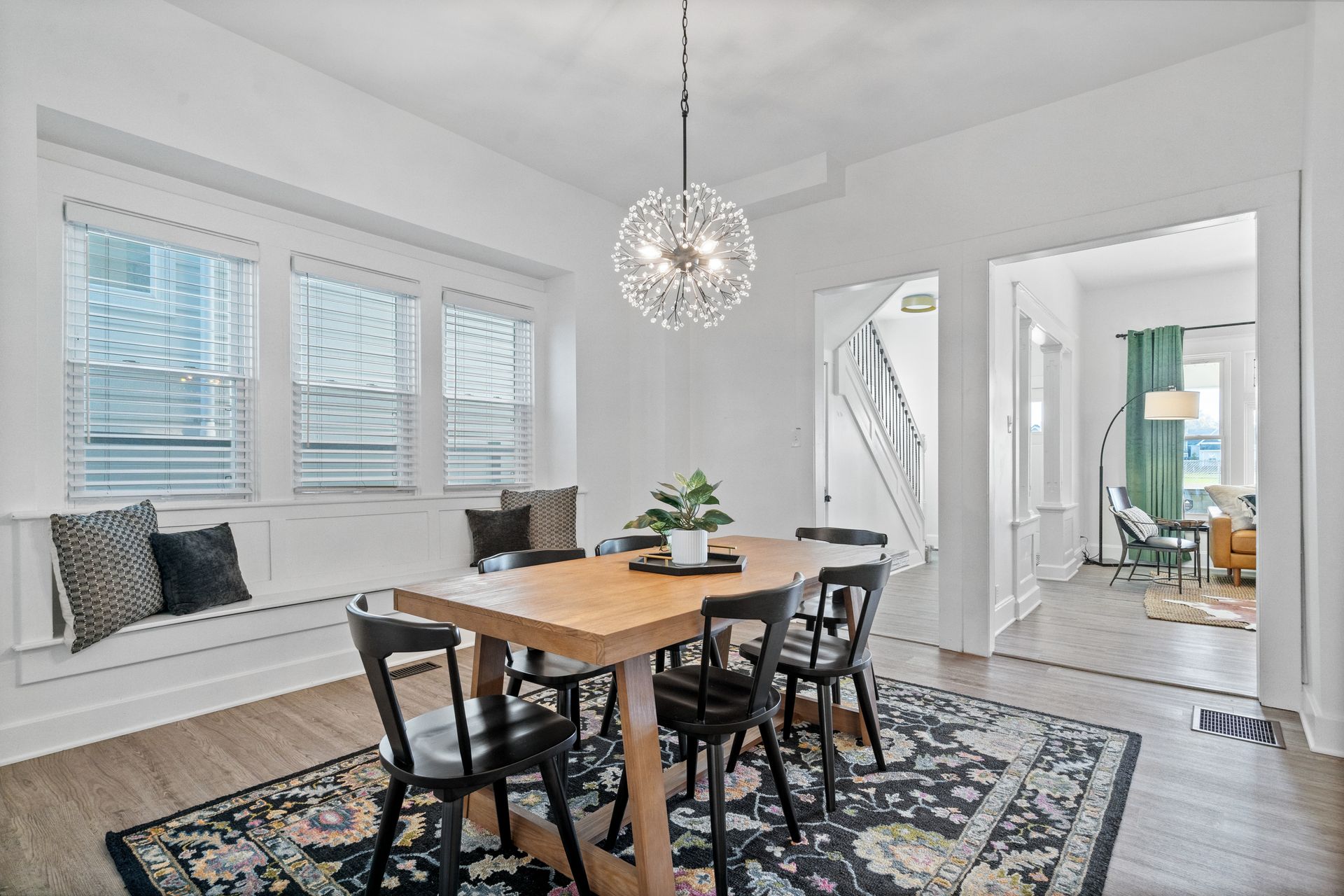 Dining room with wooden table, black chairs, and a decorative rug. A window seat and a hanging chandelier complete the space.