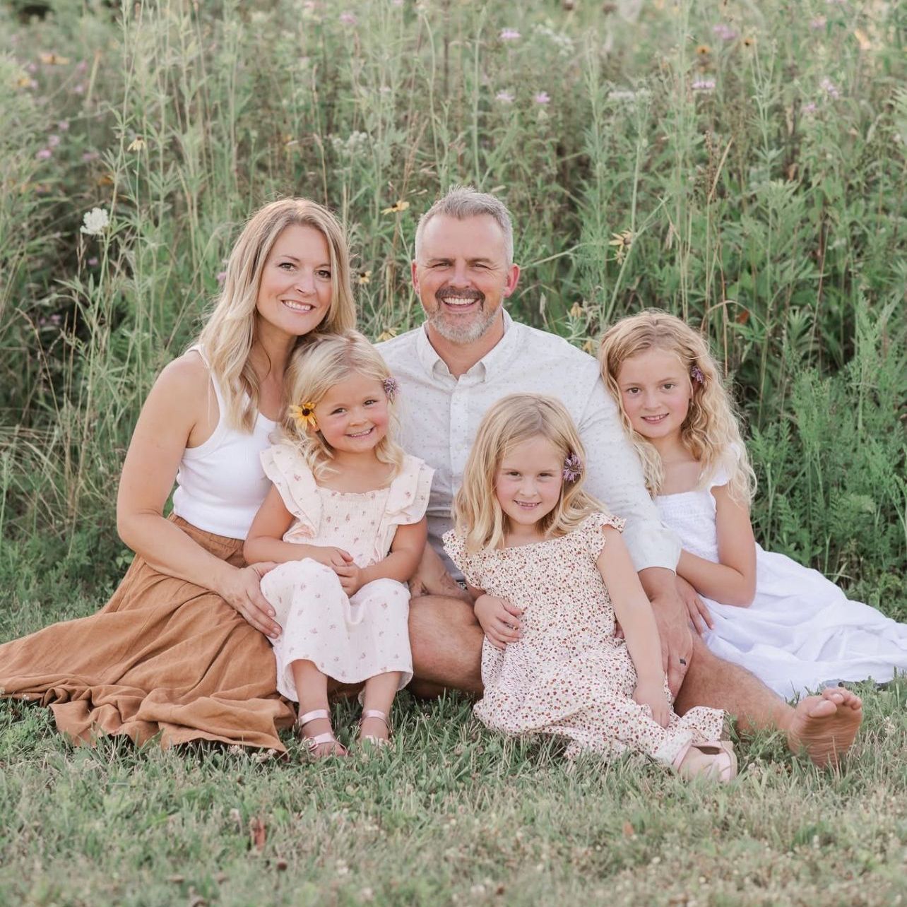 Family of five sitting in grassy field. Smiling, with tall grass in the background.