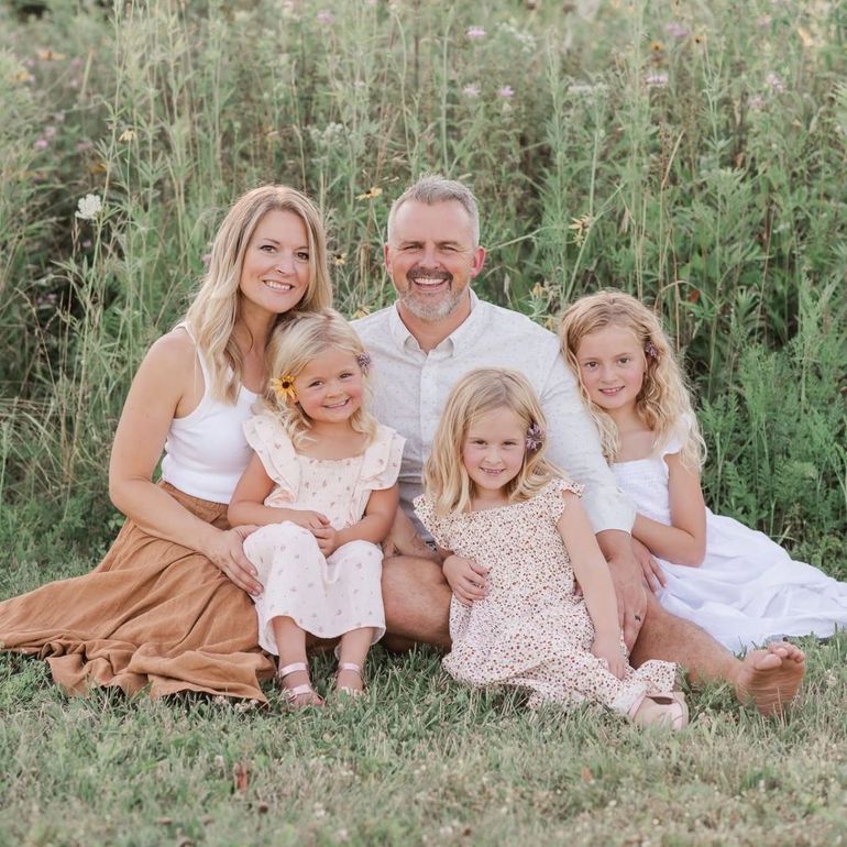 Family of five sitting in grassy field. Smiling, with tall grass in the background.