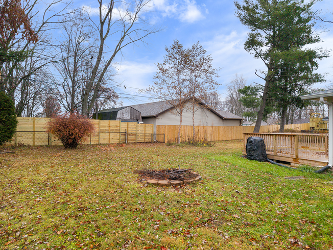 Backyard with wooden fence, fire pit, and deck, surrounded by trees and a house under a cloudy sky.