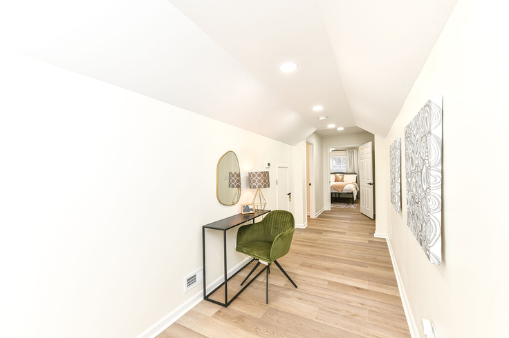 Narrow hallway with desk, chair, mirror, artwork. Light wood floors, white walls. Open doorway to a bedroom.