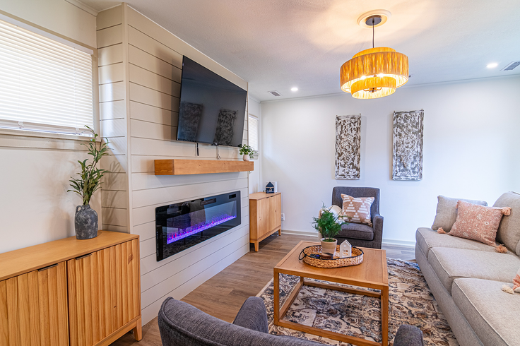 Cozy living room with a fireplace, TV, and light wood furniture. Gray couch, two armchairs, and a rug.