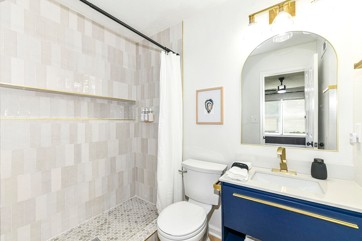 Bathroom with a blue vanity, gold accents, white toilet, and patterned shower tiles.