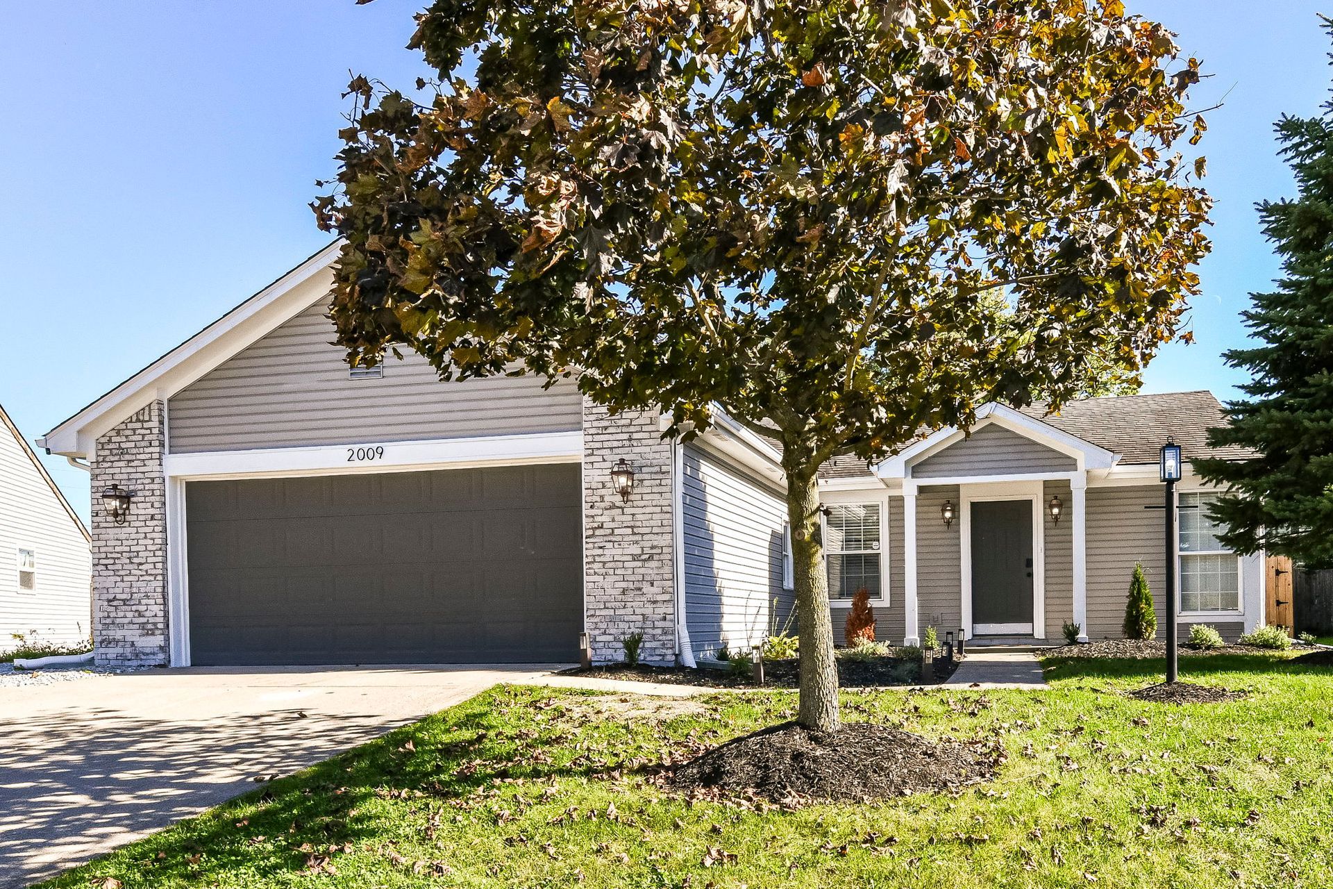 Gray house with a two-car garage, tree in front, and green lawn on a sunny day.