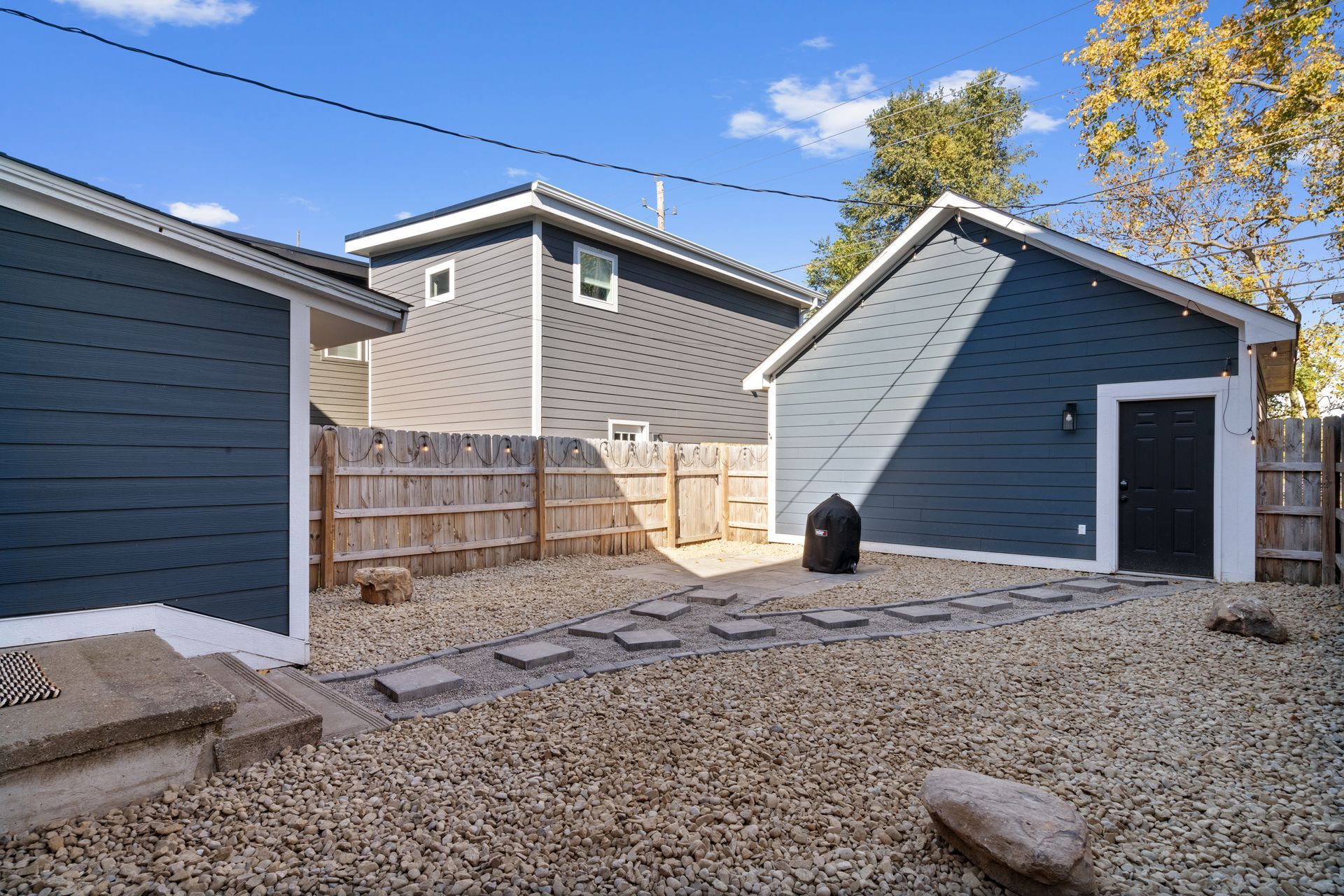 Backyard with blue buildings, gravel ground, stone path, and wooden fence on a sunny day.