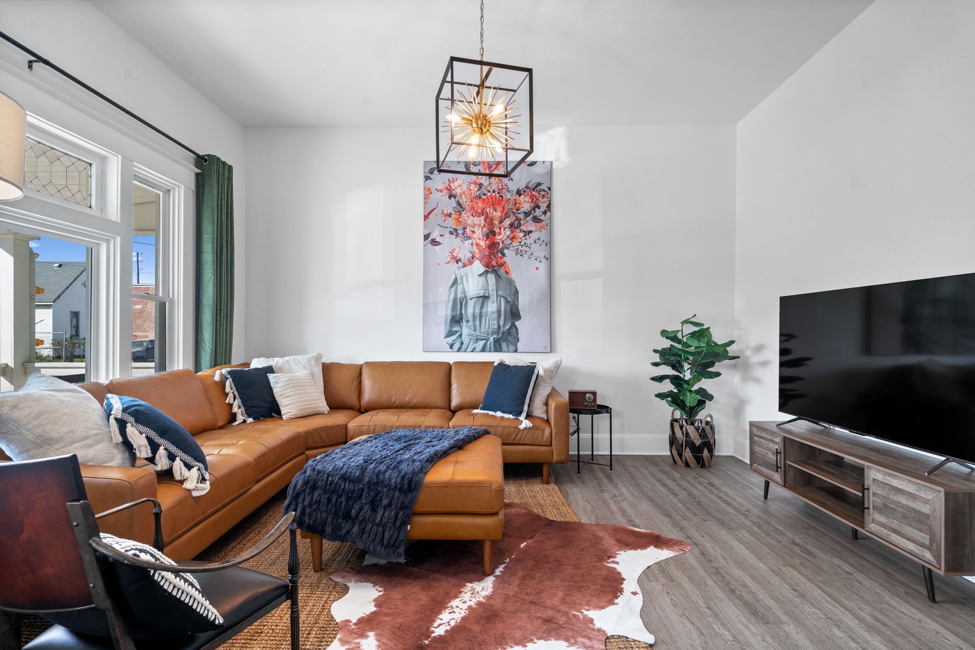 Living room with brown leather sectional, abstract art, rug, TV, and decorative chandelier.