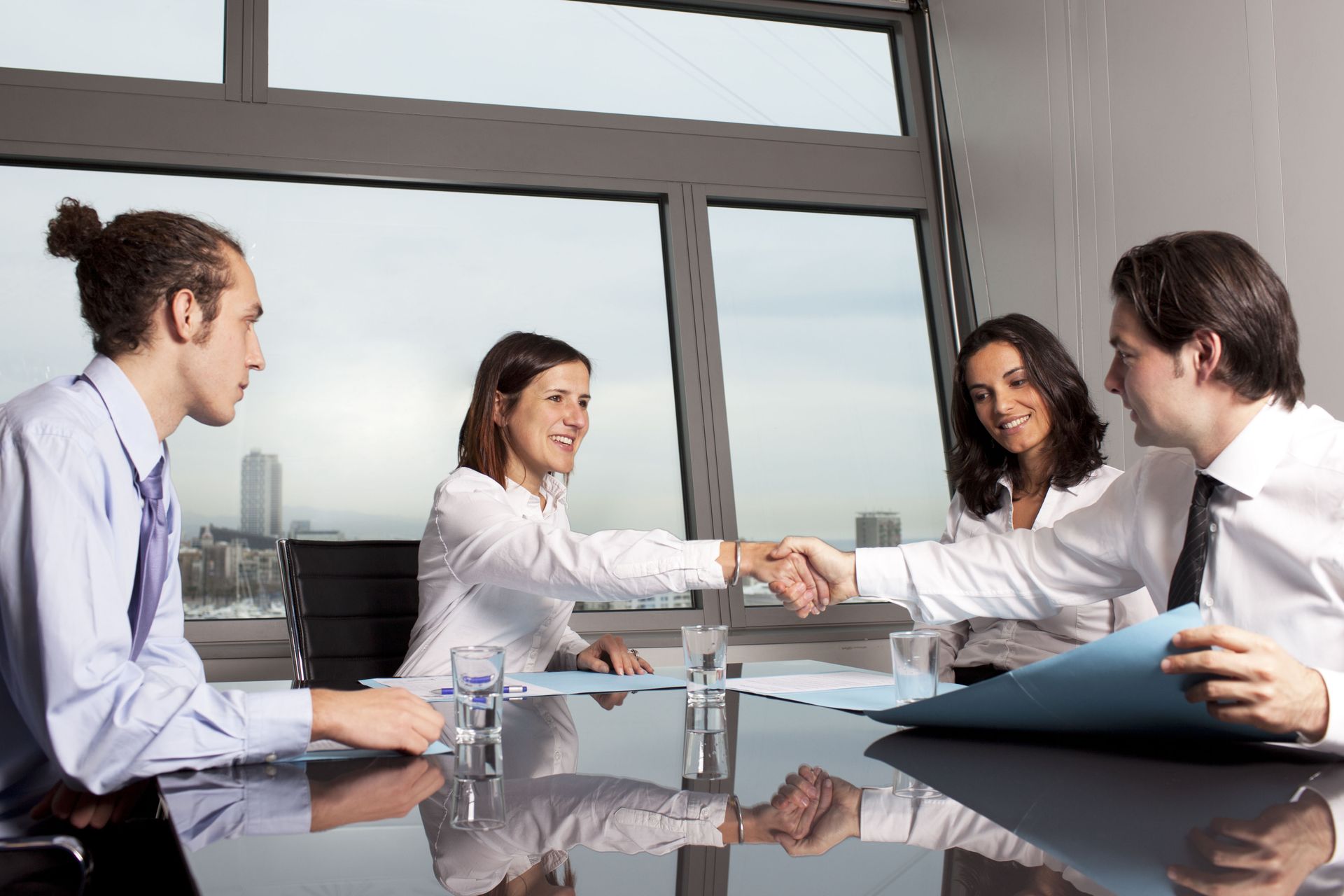 Four people at a table, two shaking hands, in front of a window overlooking a cityscape.