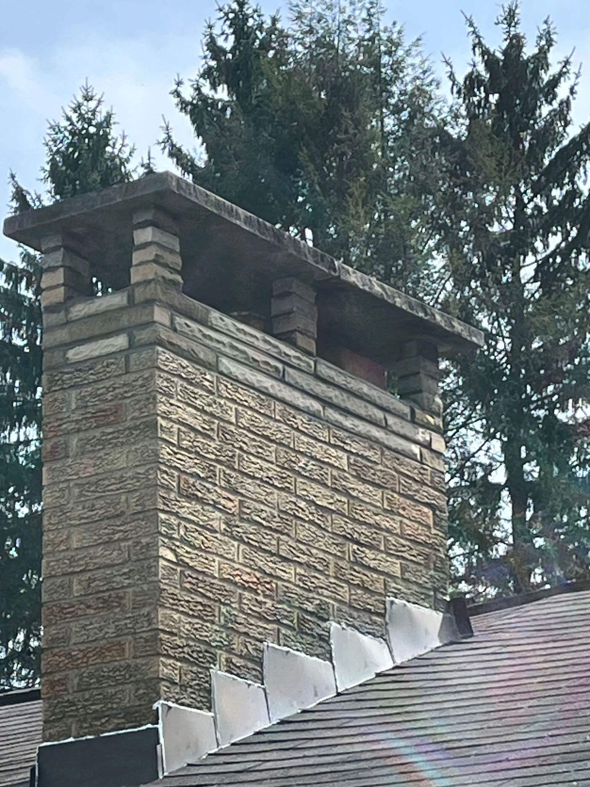Stone chimney on a rooftop with a concrete cap. Trees in the background.