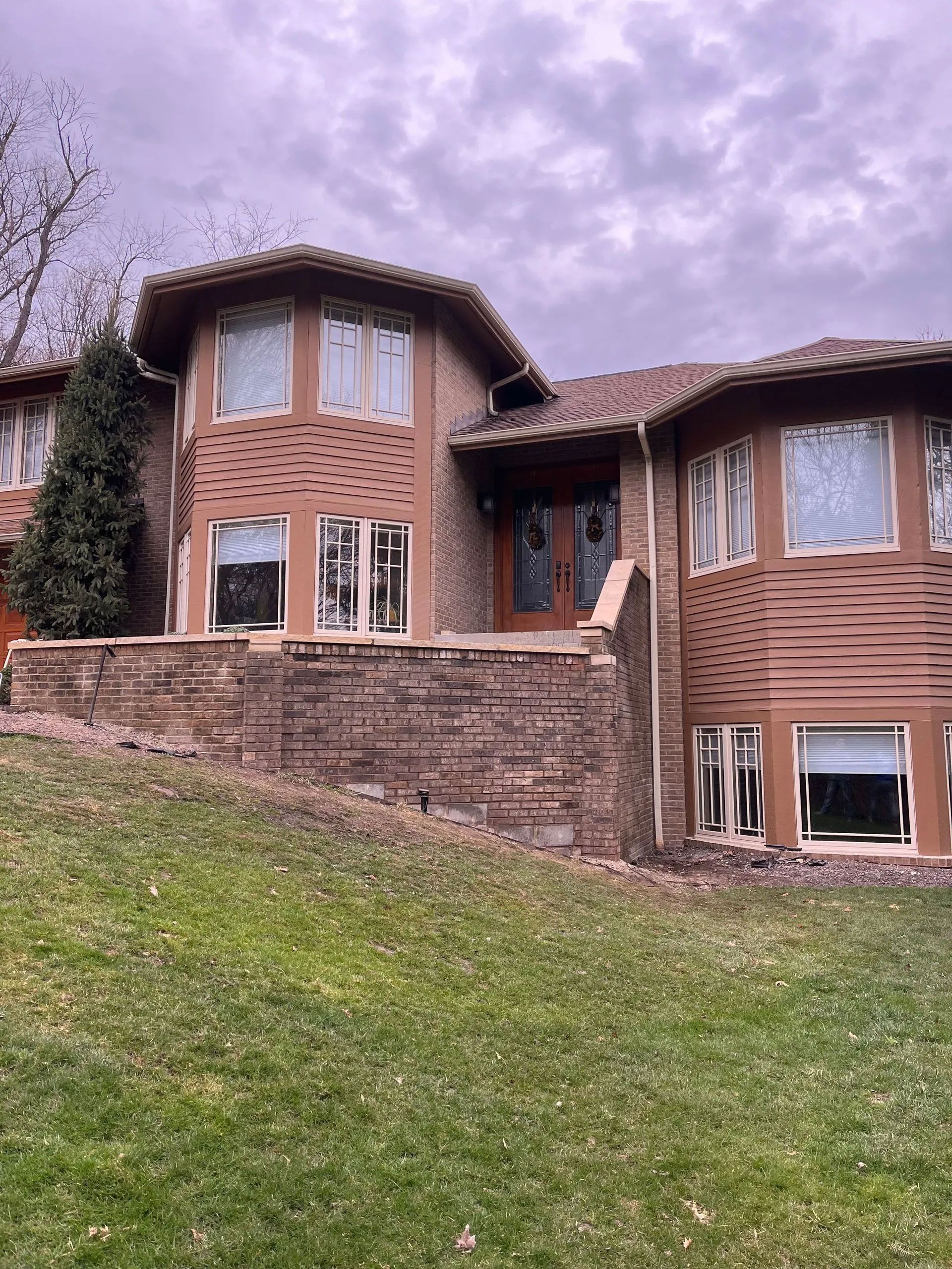 Two-story house with brown siding, stone facade, multiple windows, and a grassy lawn under a cloudy sky.
