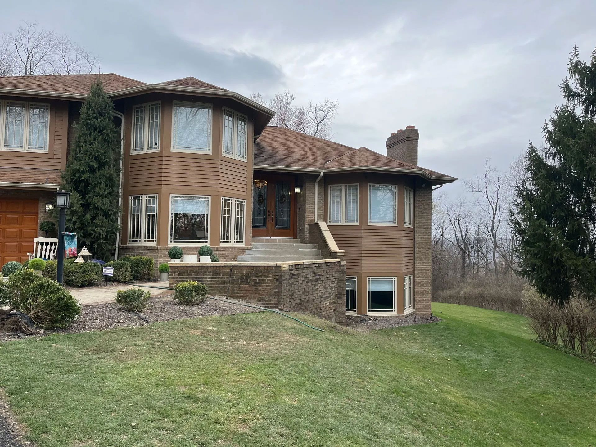 Brown brick house with bay windows and a stone wall, on a grassy hill under a cloudy sky.