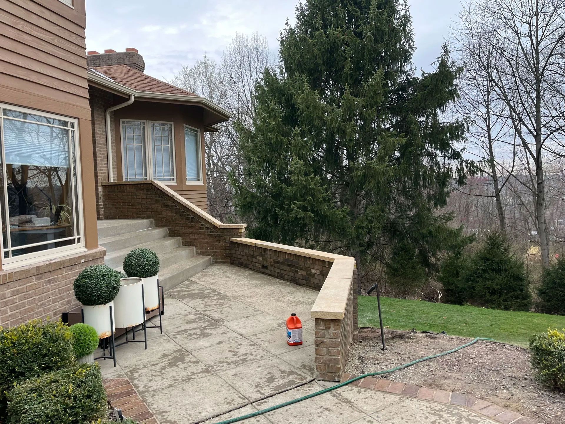 Patio with steps leading to a house. Brick walls, cement ground, and large evergreen tree in the background.